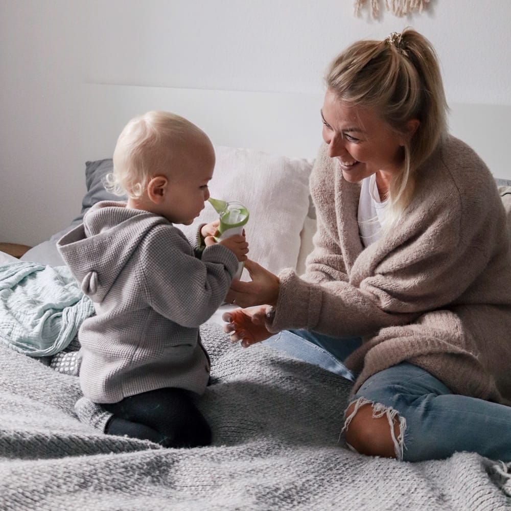 Mom and her son sitting on a bed aspirating the child's nose with a Nosiboo Go portable electric nasal aspirator